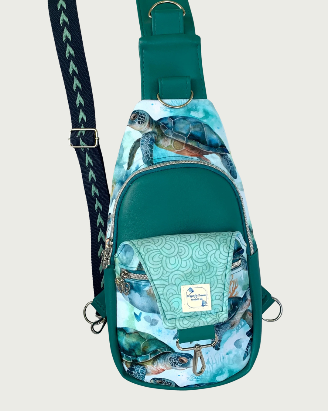 Handcrafted sling bag with a print of sweet sea turtles in teal, aqua, green, brown, and white. Water-resistant canvas bag with teal faux leather accents. It has a wide cotton canvas adjustable and repositional strap in a navy and aqua arrow pattern. The front panel lies open revealing an aqua lining with a curly print. A Dragonfly Dreams label is sewn into the lining. The main compartment of teal faux leather vinyl is zipped closed with teal zipper tape and nickel teeth.