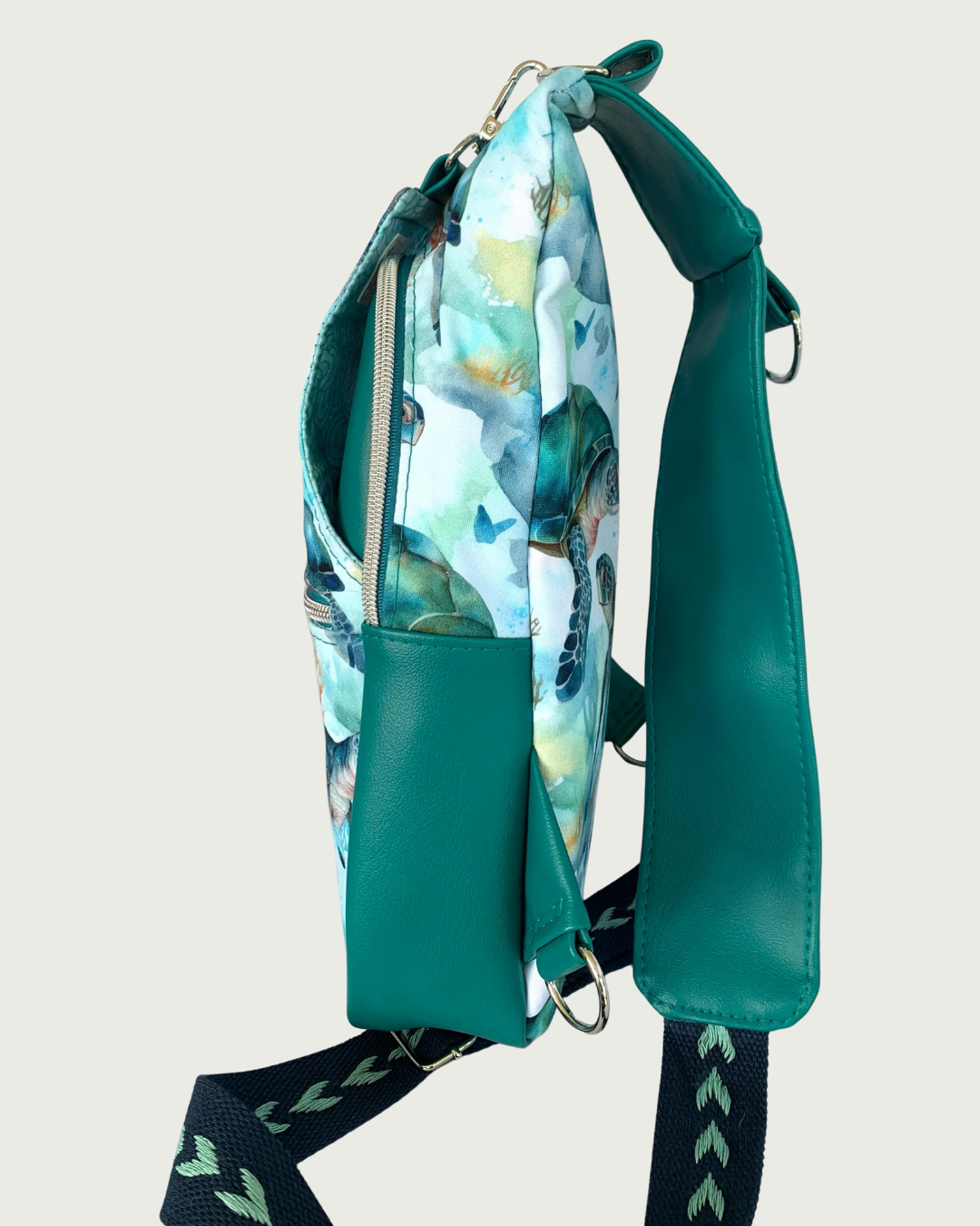 Handcrafted sling bag standing sideways with a print of sweet sea turtles in teal, aqua, green, brown, and white. Water-resistant canvas bag with teal faux leather accents with an adjustable navy and aqua wide cotton webbing strap also repositional for left and right handed wear. All hardware is nickel including the zipper teeth on teal zipper tape. A front panel that unhooks with a swivel held onto a d-ring is shown. Behind that is a curved zippered main compartment of teal vinyl.