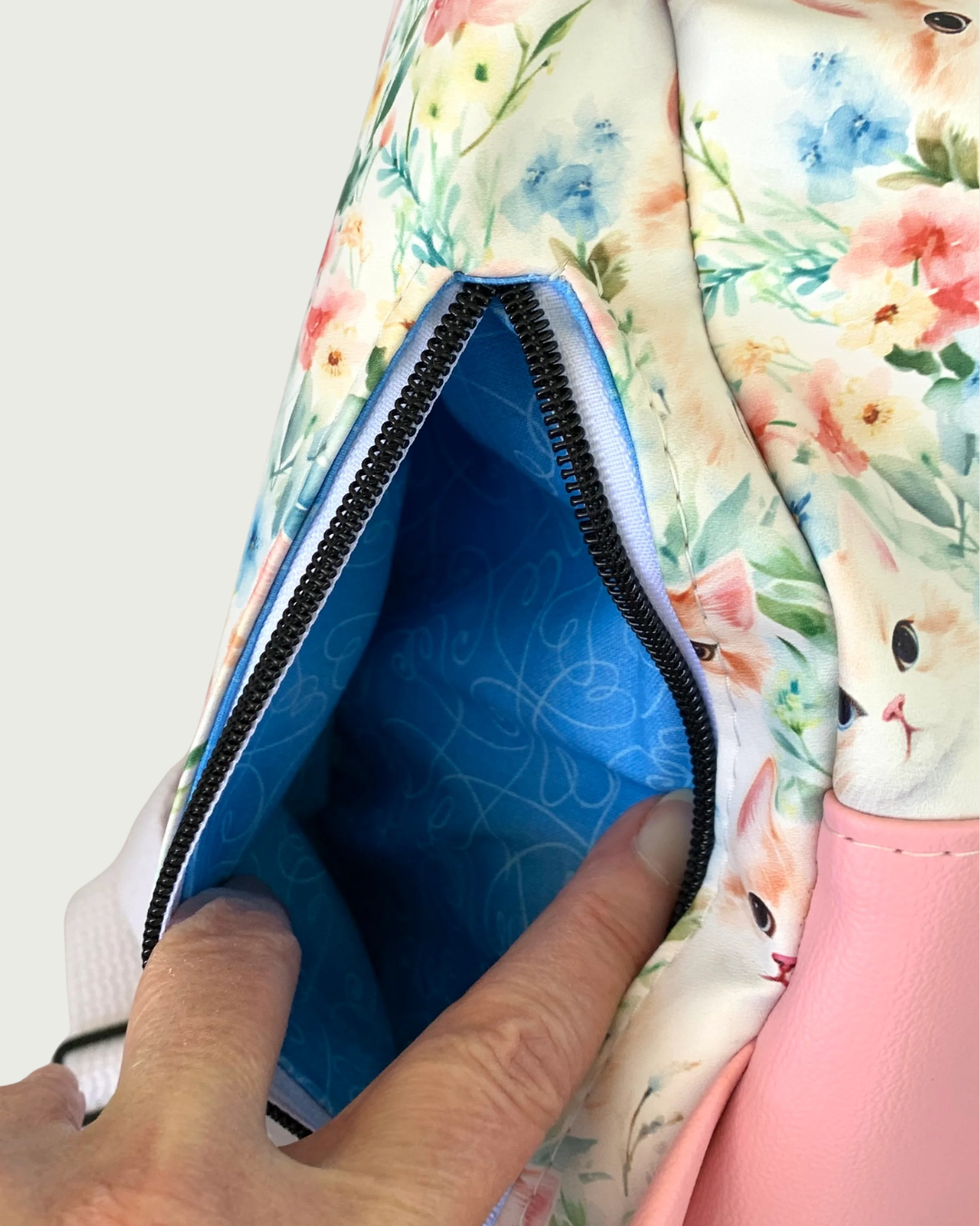 Handcrafted Sling bag with vintage kitty faces and a pink vinyl gusset. A hand is holding open the back vertical zipper revealing a blue lining with white swirling hearts. The zipper teeth are matte black on white zipper tape.