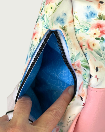 Handcrafted Sling bag with vintage kitty faces and a pink vinyl gusset. A hand is holding open the back vertical zipper revealing a blue lining with white swirling hearts. The zipper teeth are matte black on white zipper tape.