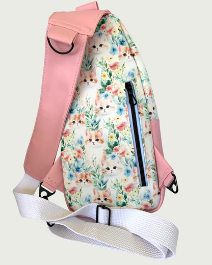 The back side of a handcrafted sling bag with vintage kitty faces featuring pink vinyl accents including a wide shoulder strap with an adjustable white cotton webbing strap extension also repositional for left and right handed wear. All hardware is matte black including the vertical zipper teeth on white zipper tape. A large D-ring is attached to the shoulder strap for easy hanging.