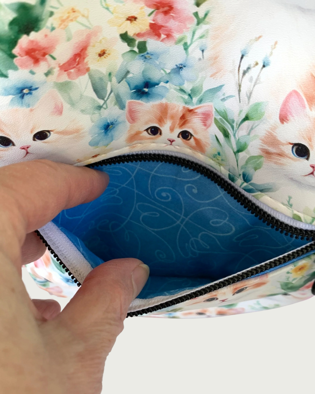 Handcrafted Sling bag with vintage kitten faces closeup view of fingers holding open a front zippered pocket revealing a blue lining with white swirly hearts. The zipper teeth are matte black on white zipper tape.