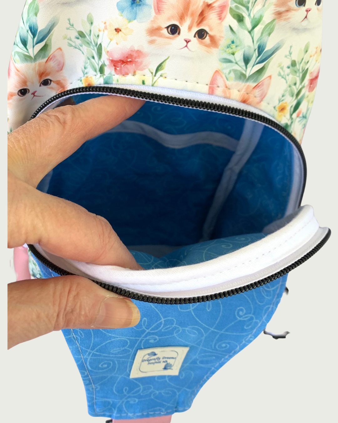 Main compartment of handcrafted sling bag with vintage kitty faces. The lining of the compartment is blue with white swirly hearts and a Dragonfly Dreams label sewn into the inside of the front panel. A slip pocket appears inside as well as white-bound seams. The zipper teeth are matte black on white zipper tape.