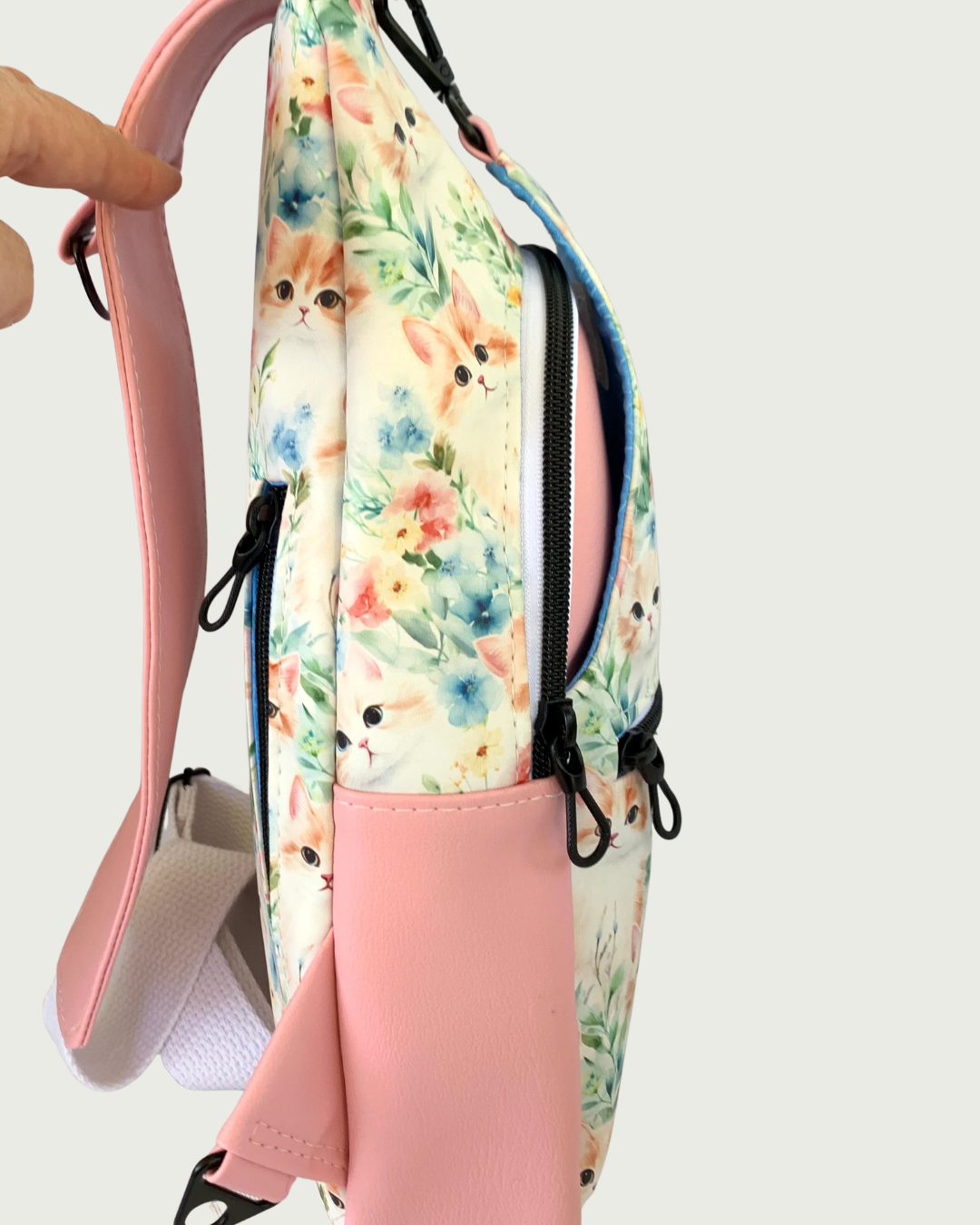 The left side view of a handcrafted sling bag with vintage kitty faces featuring pink vinyl accents including a wide shoulder strap with an adjustable white cotton webbing strap extension also repositional for left and right handed wear. All hardware is matte black including the zipper teeth and zipper pulls.