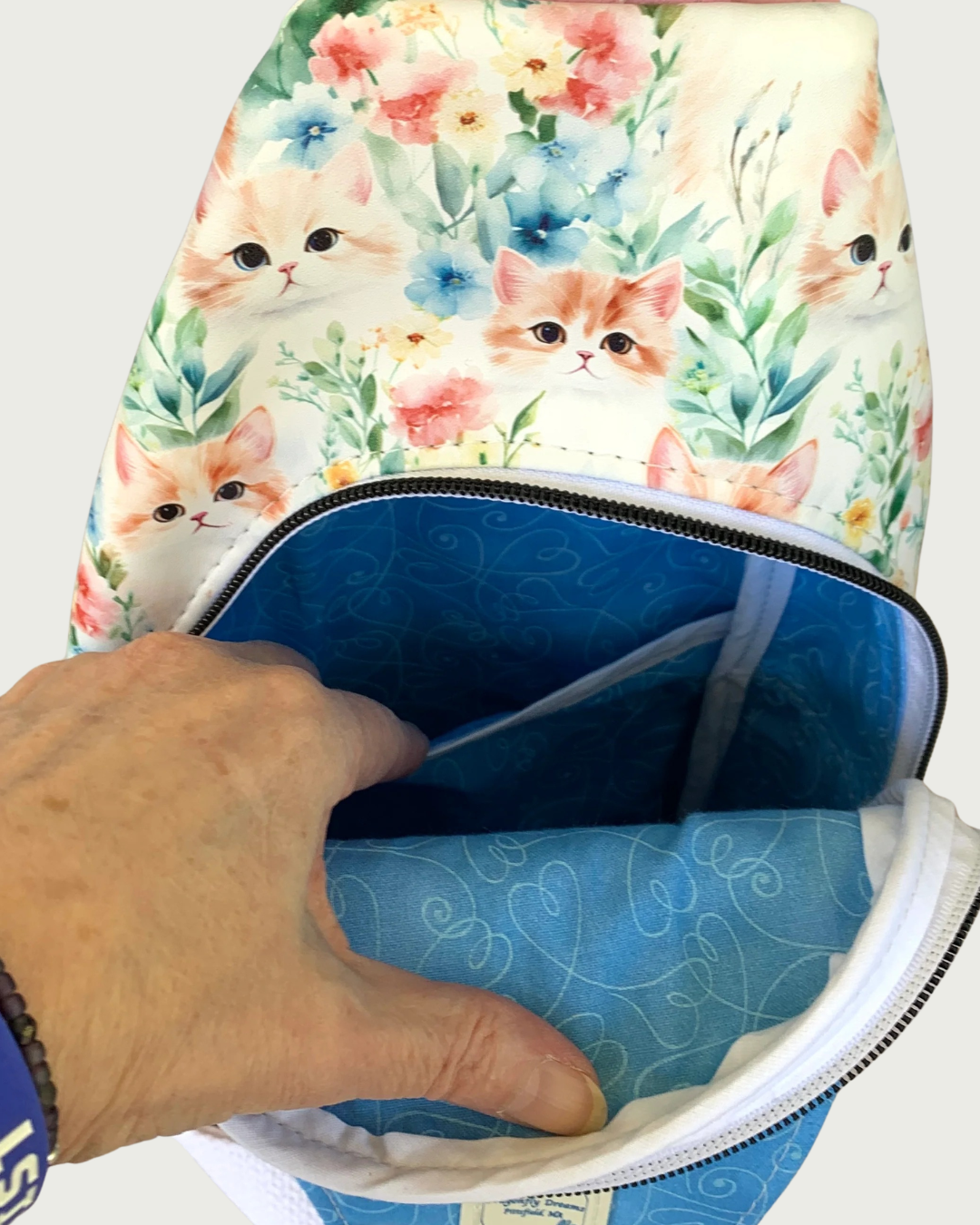 Main compartment of handcrafted sling bag with vintage kitty faces. The lining of the compartment is blue with white swirly hearts and a Dragonfly Dreams label sewn into the inside of the front panel. A hand is holding open a slip pocket. The zipper teeth are matte black on white zipper tape.