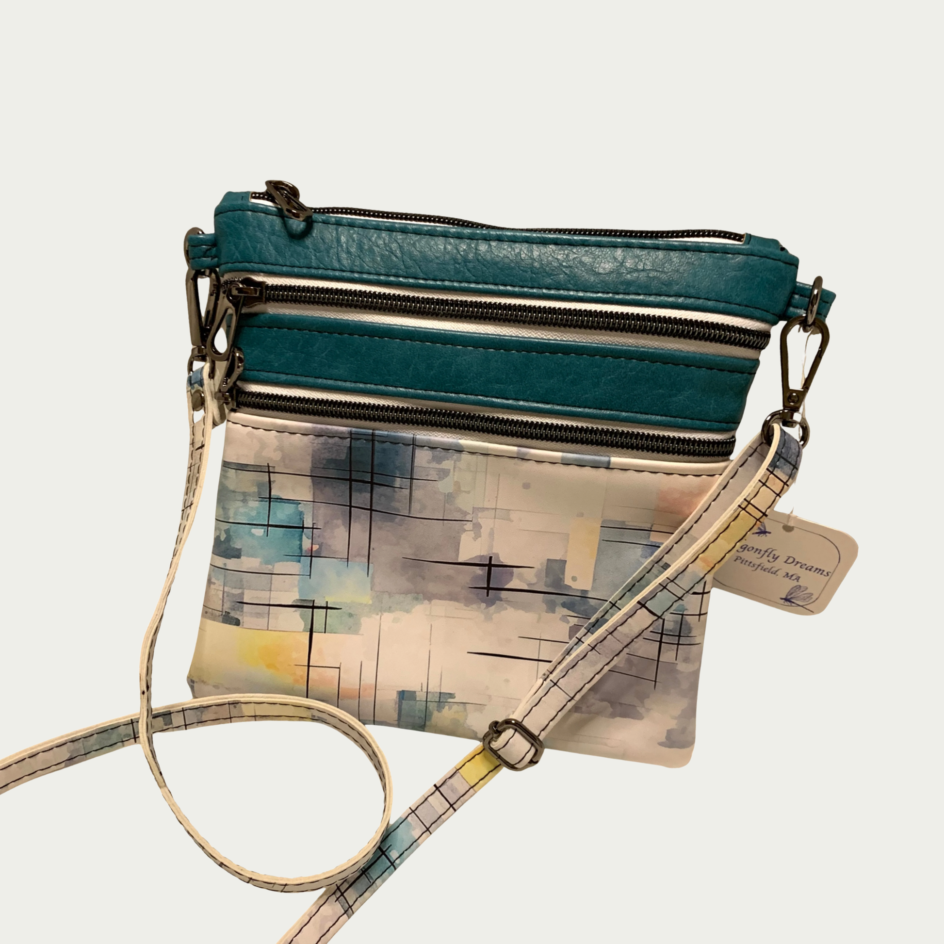 A crossbody bag with multiple zippered pockets made from teal faux leather and a watercolored urban printed vinyl. The detachable strap is made from the same printed material. Matt black hardware.