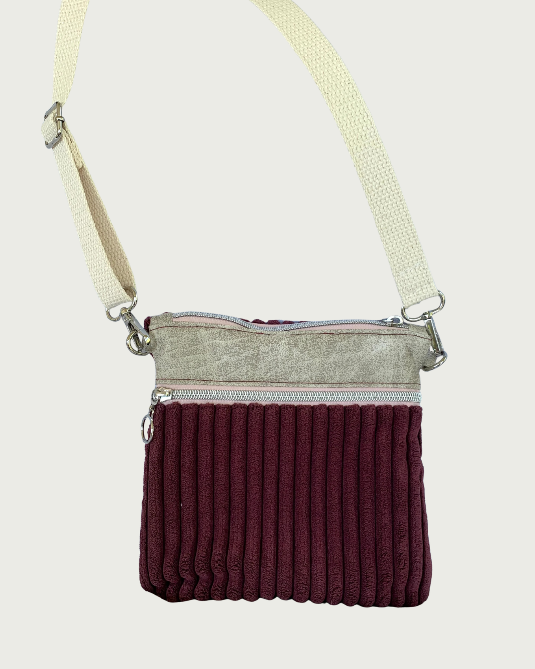 Handmade maroon wide-wale corduroy crossbody bag with sand-colored distressed vinyl accents hanging by an adjustable and detachable cotton webbing, natural colored strap. Nickel hardware throughout. There is one pocket with pink zipper tape as well as a top zippered closure. 