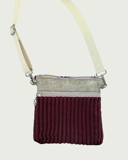 Handmade maroon wide-wale corduroy crossbody bag with sand-colored distressed vinyl accents hanging by an adjustable and detachable cotton webbing, natural colored strap. Nickel hardware throughout. There is one pocket with pink zipper tape as well as a top zippered closure. 