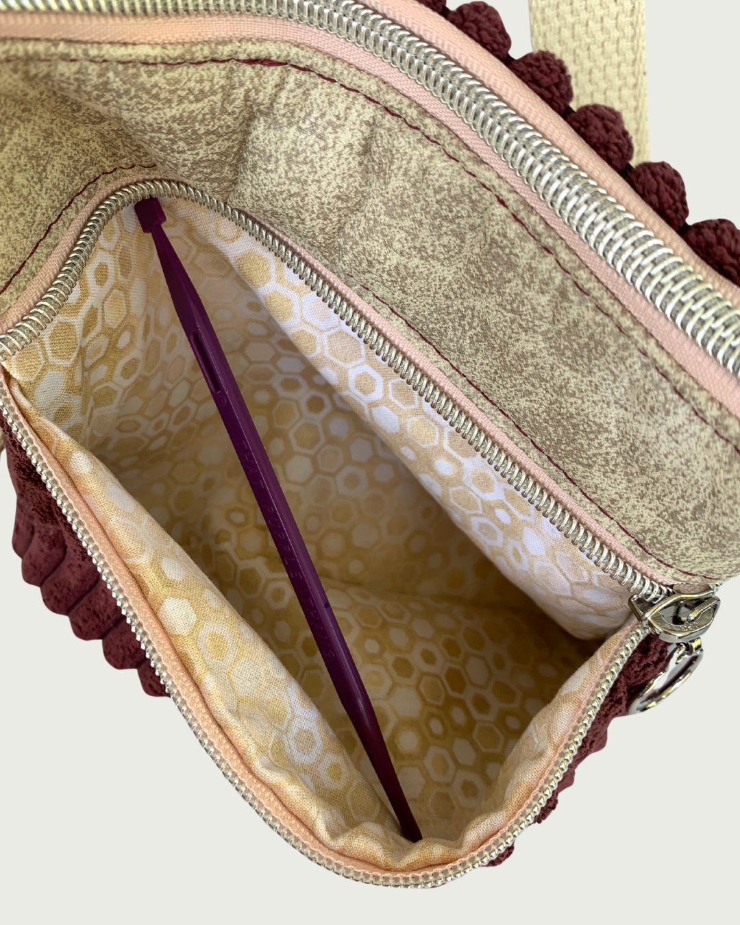 Top view of a handmade maroon wide-wale corduroy crossbody bag with sand-colored distressed vinyl accents. The top zipper closure is pink with nickel zipper as is the open zippered pocket exposing a small geometric print of tan, gold, and white lining. This pocket is held open with a purple stick.
