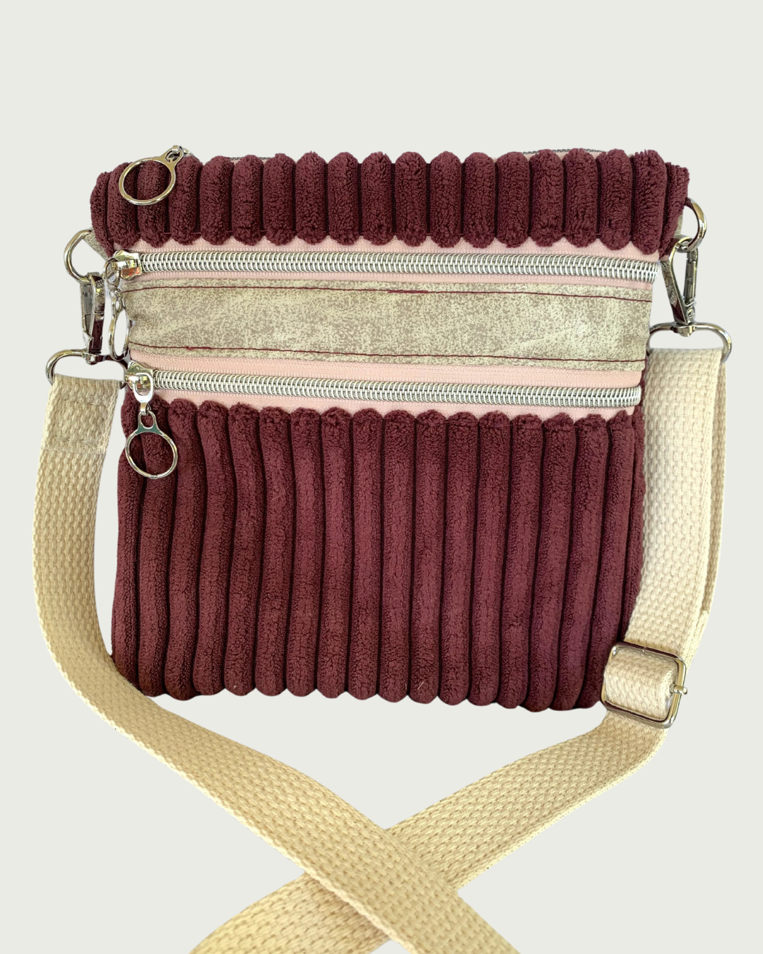 Close view of a handmade maroon wide-wale corduroy crossbody bag with sand-colored distressed vinyl accents. Attached is an adjustable and detachable cotton webbing, natural colored strap. Nickel hardware throughout. There are two pockets with pink zipper tape. 