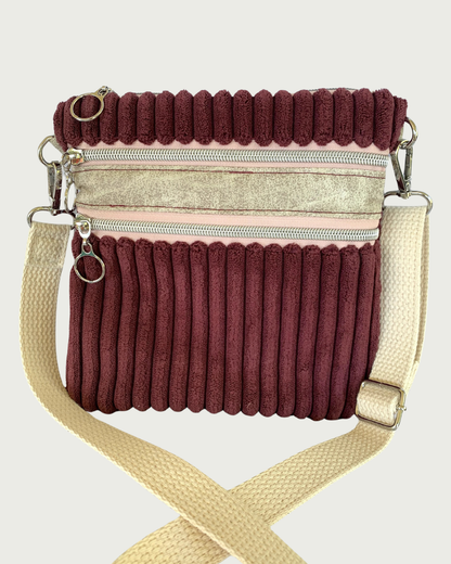 Close view of a handmade maroon wide-wale corduroy crossbody bag with sand-colored distressed vinyl accents. Attached is an adjustable and detachable cotton webbing, natural colored strap. Nickel hardware throughout. There are two pockets with pink zipper tape. 