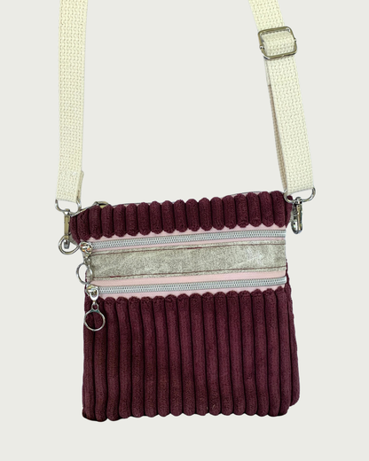 Handmade maroon wide-wale corduroy crossbody bag with sand-colored distressed vinyl accents hanging by an adjustable and detachable cotton webbing, natural colored strap. Nickel hardware throughout. There are two pockets with pink zipper tape. 