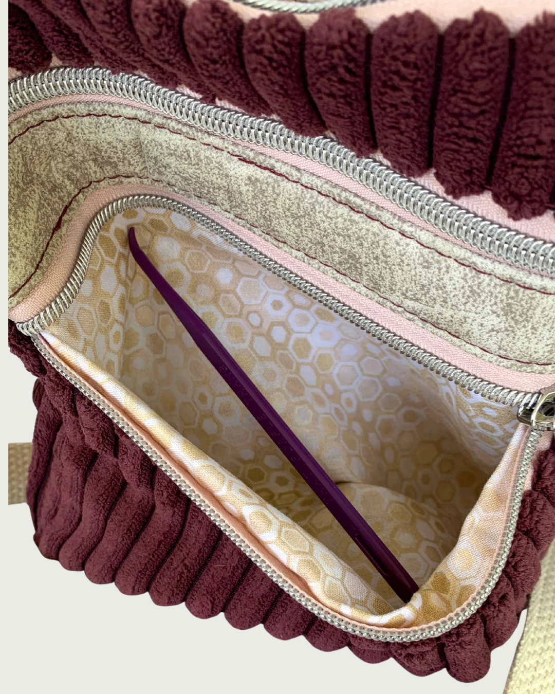 Top view of a handmade maroon wide-wale corduroy crossbody bag with sand-colored distressed vinyl accents. The top zipper closure is pink with nickel zipper as are the two zippered pockets. The lower pocket is open exposing a small geometric print of tan, gold, and white lining. This pocket is held open with a purple stick.