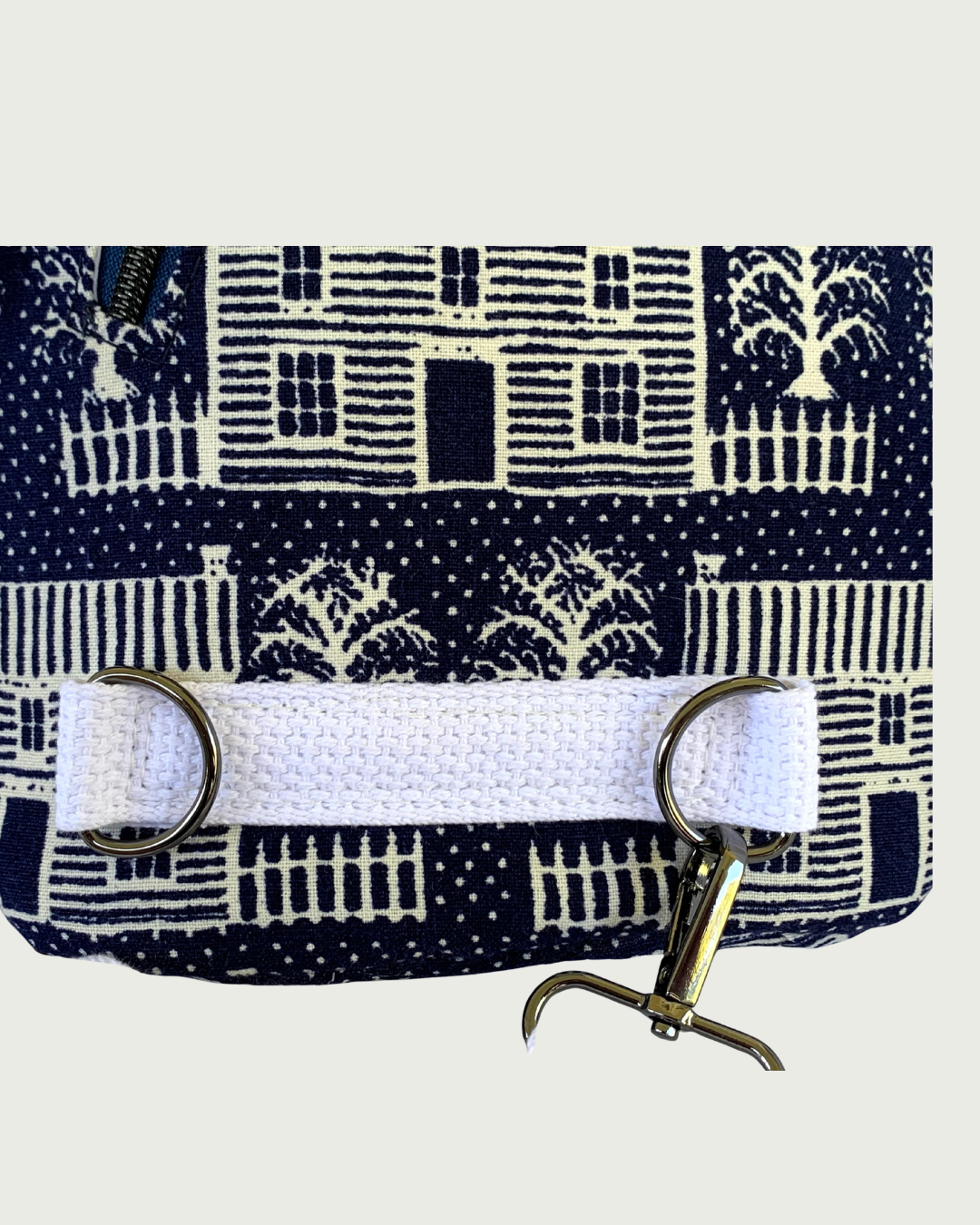 Handcrafted navy crossbody sling bag with off-white houses closeup of sewn-in cotton webbing strap securing 2 d-rings. The gunmetal swivel clasp from an adjustable strap is attached to one d-ring.