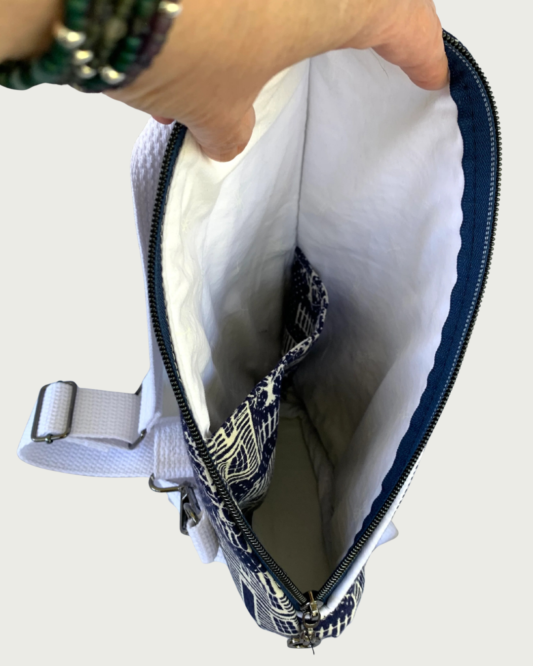 Handmade crossbody sling bag of navy and off-white houses and picket fences with a hand holding open a navy zipper exposing the bag interior of white lining material and the same navy and white print on a slip pocket. The bag interior looks very spacious. Also showing is a white adjustable cotton webbing strap and gunmetal hardware.