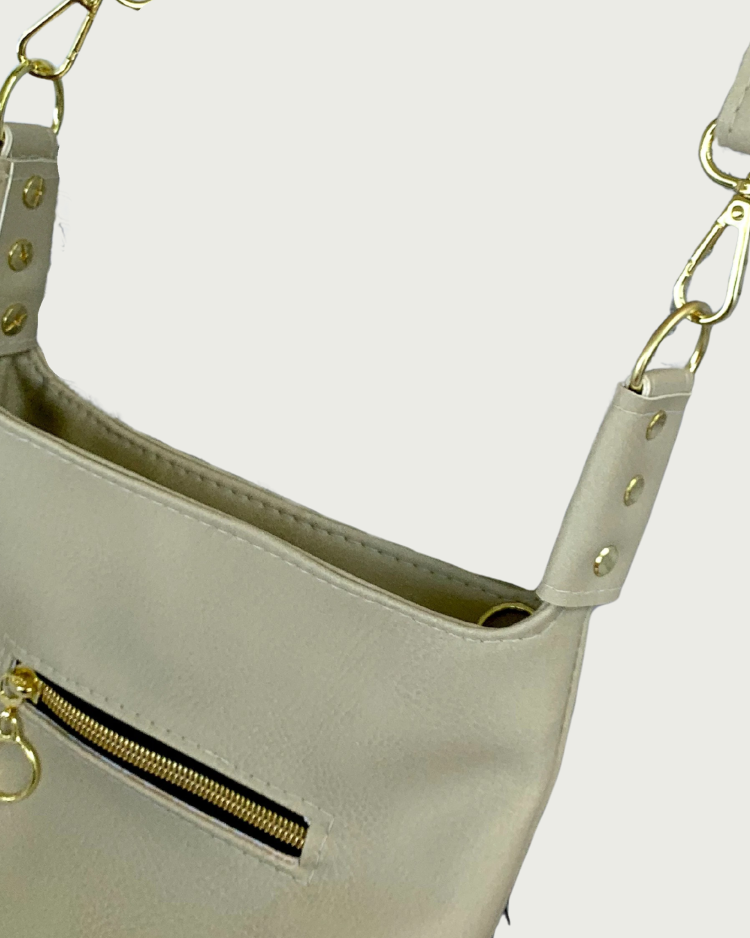 Close-up of a light beige shoulder bag with gold-tone hardware, a zippered pocket, and a strap connector with gold rivets.