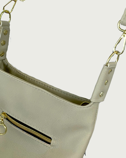 Close-up of a light beige shoulder bag with gold-tone hardware, a zippered pocket, and a strap connector with gold rivets.